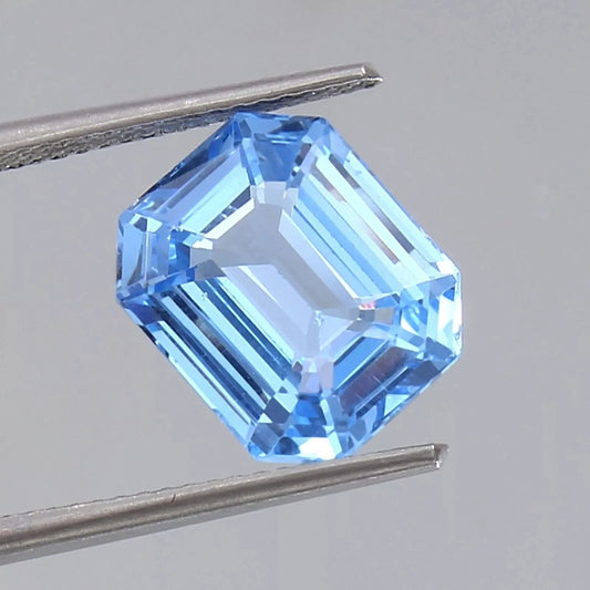 Blue Spinel gemstone held between tweezers on a gray background