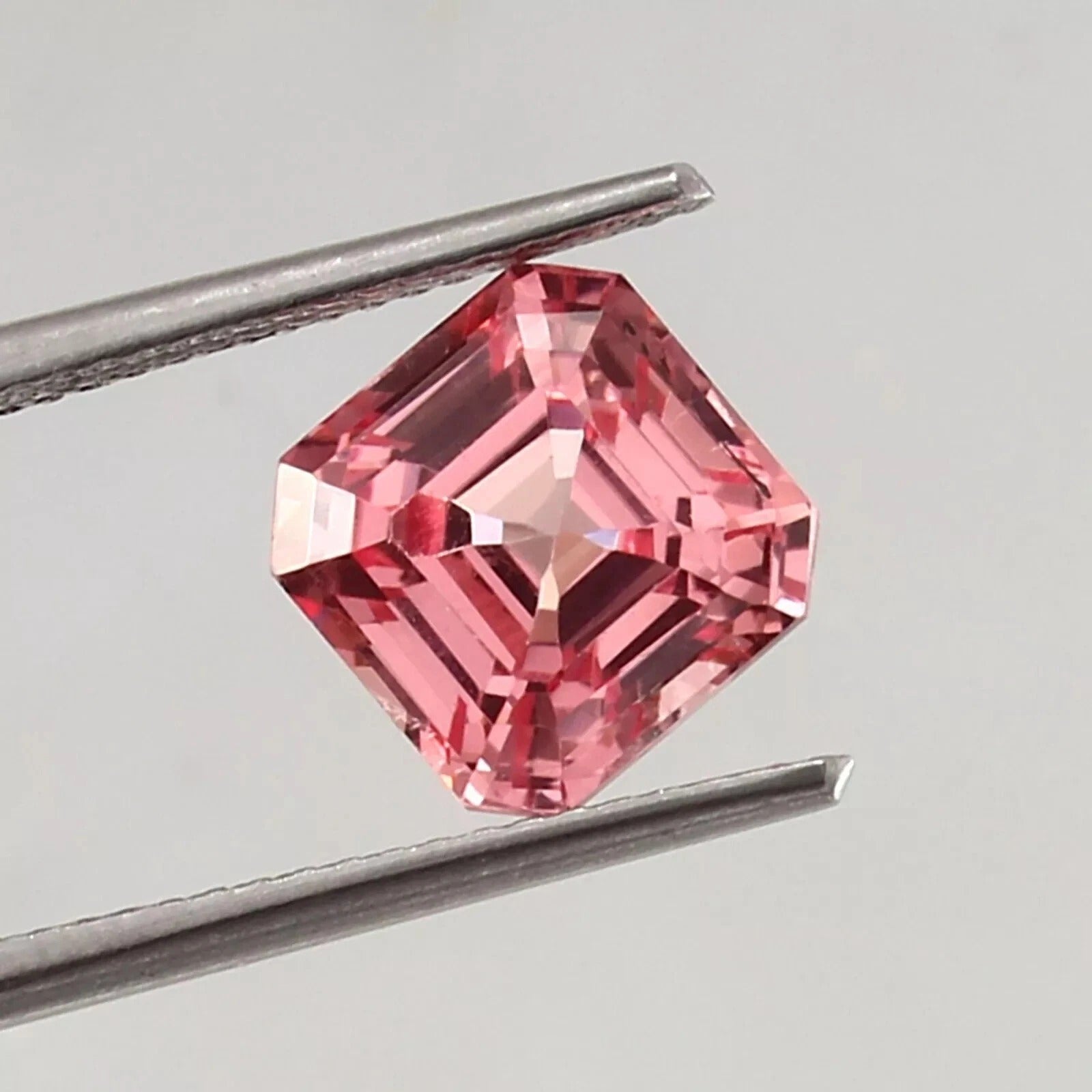 Natural Padparadscha Sapphire gemstone held by tweezers on a gray background