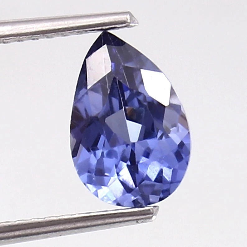 Teardrop-shaped blue Kashmir Sapphire gemstone held by tweezers on a white background