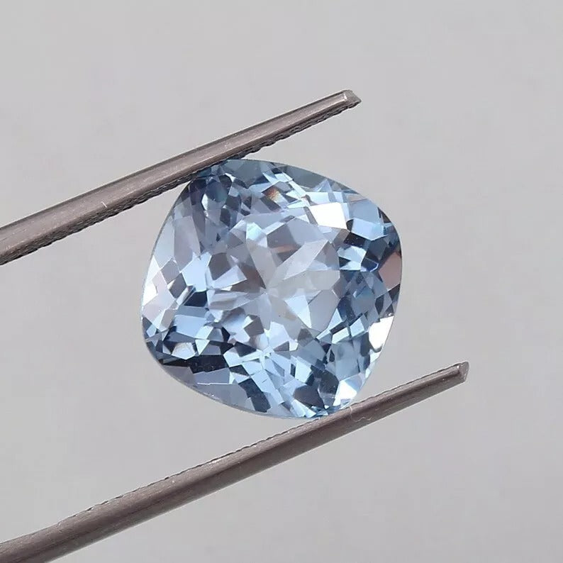 Blue Aquamarine gemstone held between tweezers against a neutral background