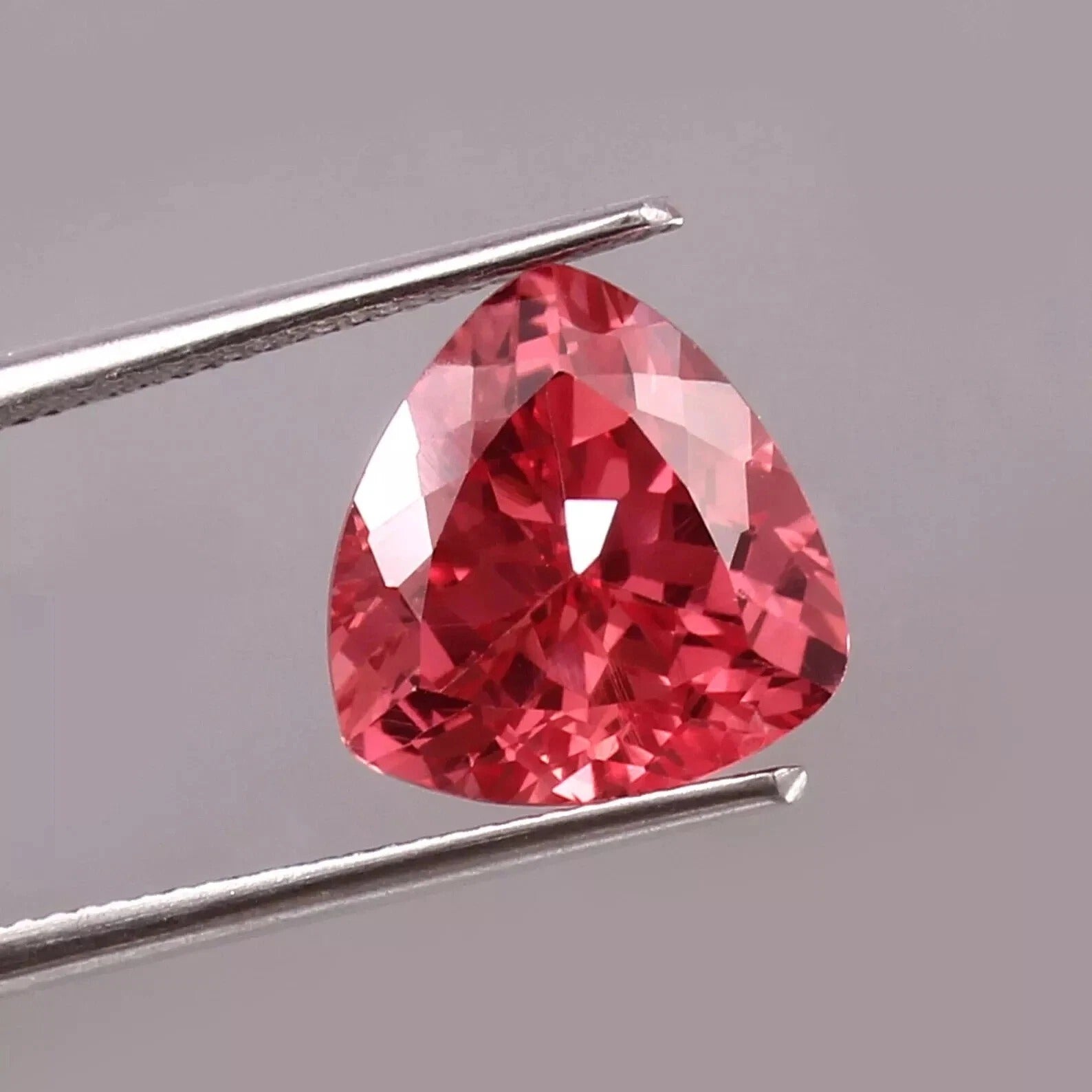 Trillion-shaped Padparadscha Sapphire gemstone held by tweezers on a gray background