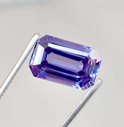 Faceted Alexandrite gemstone held between two metal prongs on a light gray background
