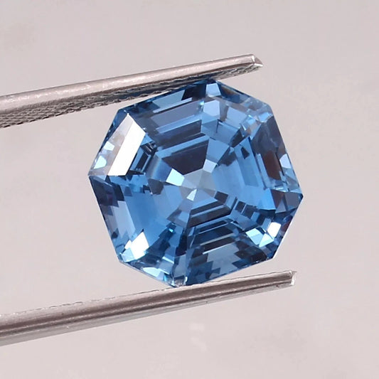 Asscher blue Aquamarine gemstone held between tweezers on a light gray background
