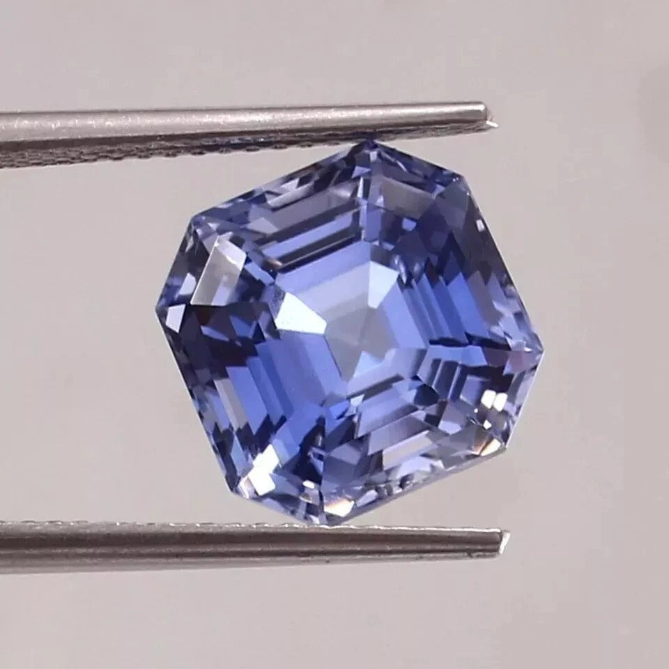 Blue sapphire gemstone held between tweezers on a light gray background