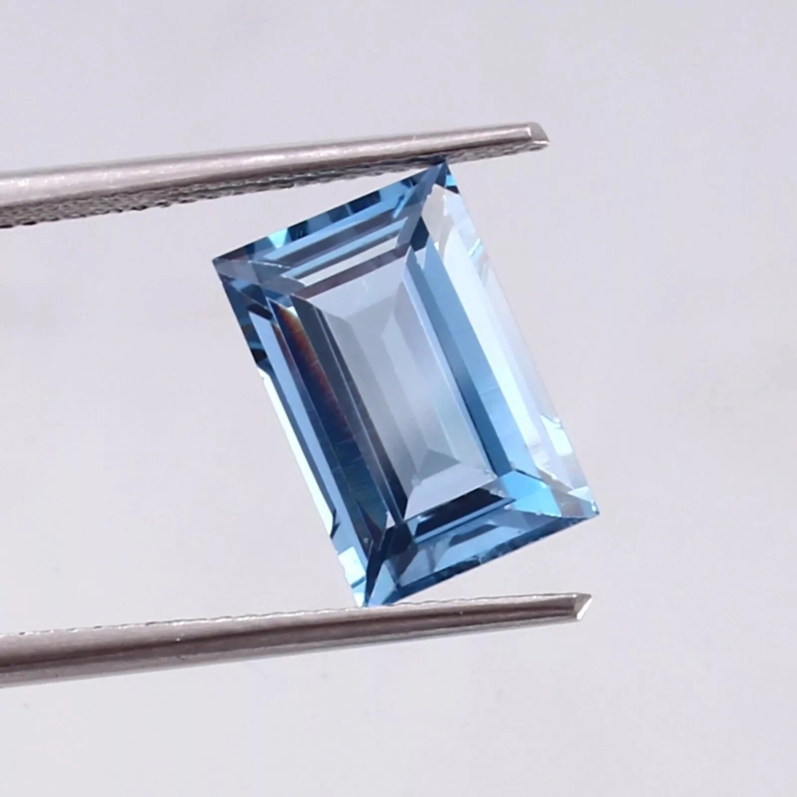 Aquamarine blue gemstone held by tweezers against a light gray background