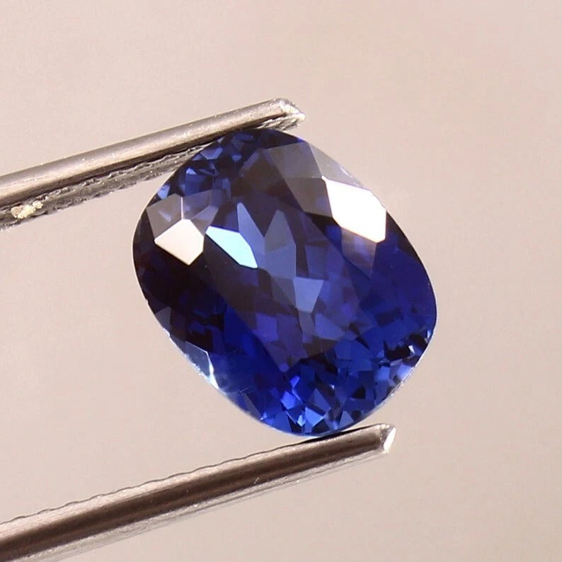 Faceted Royal Blue Sapphire gemstone held between tweezers on a beige background