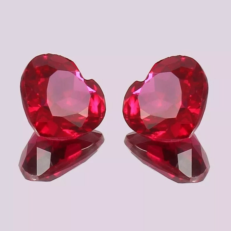 Pair of red ruby heart-shaped gemstones on a reflective surface with a light purple background