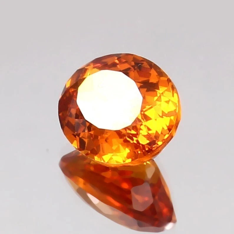 Faceted orange Sapphire gemstone on a reflective surface with a light gray background