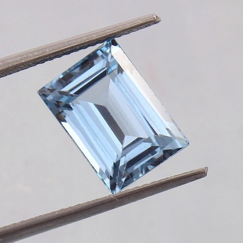 Blue Brazilian Aquamarine Loose Baguette Cut gemstone held between tweezers on a neutral background