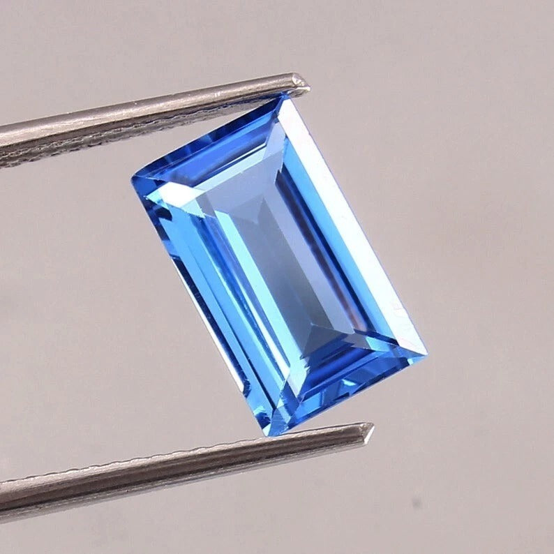 Blue Brazilian Aquamarine Baguette Cut gemstone held by tweezers on a gray background
