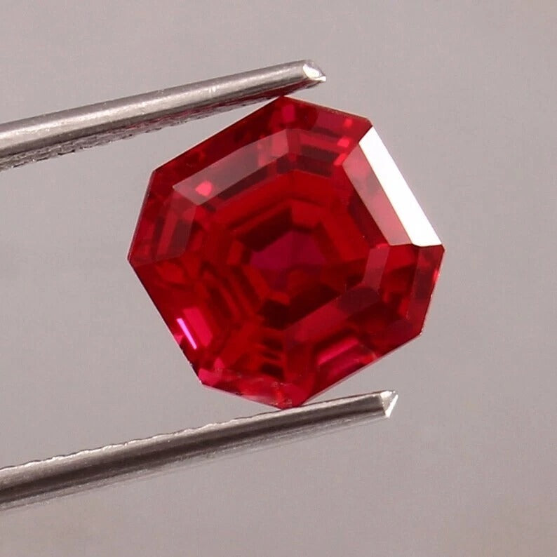Hexagonal Pigeon Blood Ruby gemstone held by tweezers on a gray background