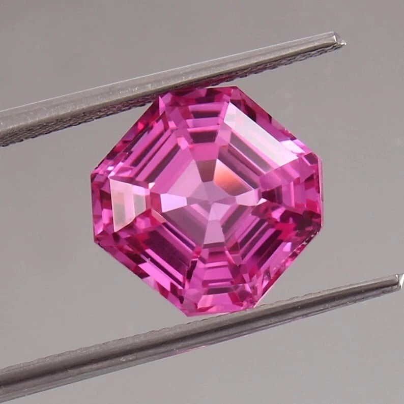 Pink Sapphire asscher gemstone held between tweezers on a gray background