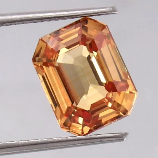 Peach Padparadscha gemstone held between tweezers on a gray background