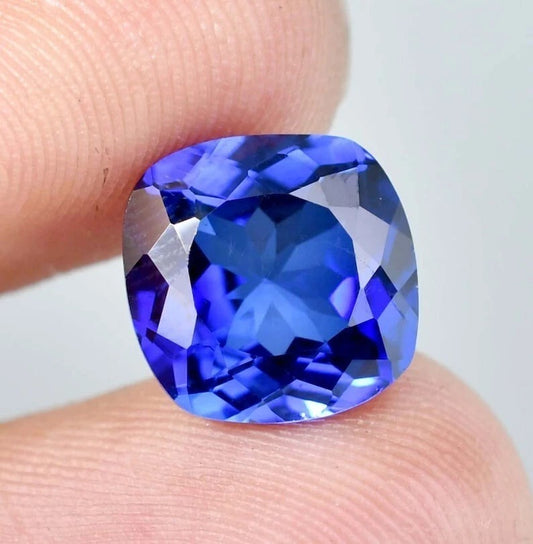 Blue Sapphire cushion gemstone held between two fingers against a neutral background
