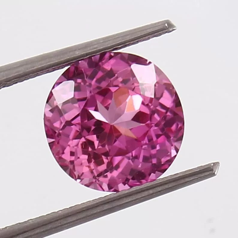 Round Pink Sapphire gemstone held between tweezers on a light gray background