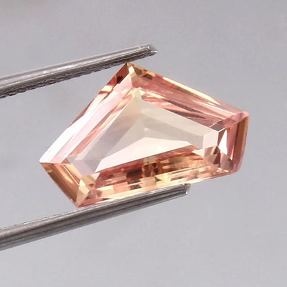 Faceted Padparadscha Sapphire gemstone held by tweezers against a gray background
