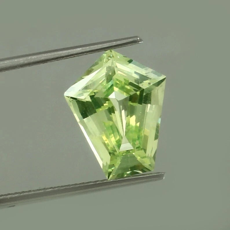 Green Chrysoberyl gemstone held by tweezers against a gray background
