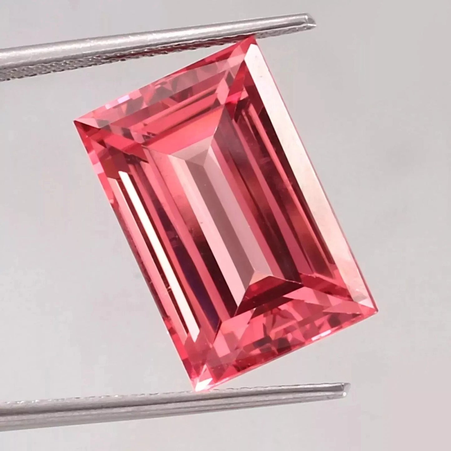 Padparadscha Sapphire – Baguette Cut gemstone held by tweezers on a light gray background