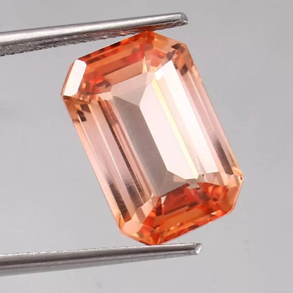 Bi-Color Padparadscha gemstone held by tweezers on a gray background