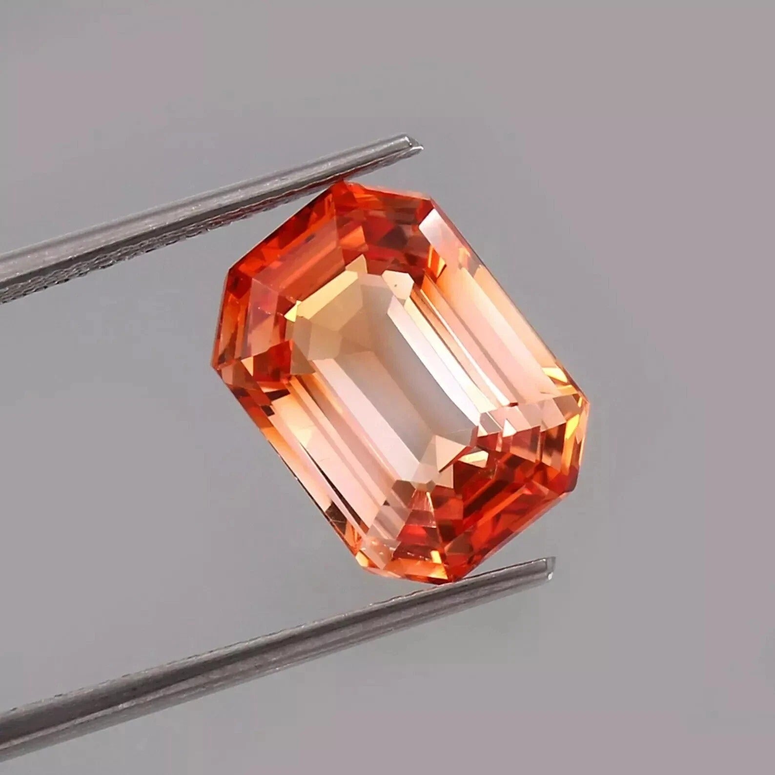 Faceted Bi-Color Unheated Padparadscha sapphire gemstone held between tweezers on a gray background