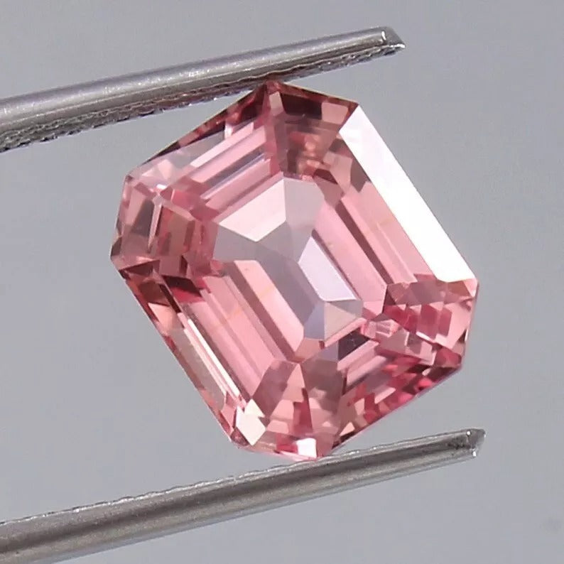 Pink Padparadscha Sapphire gemstone held between tweezers on a gray background