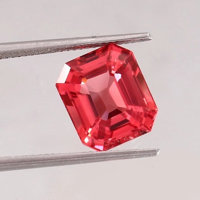 Padparadscha Sapphire gemstone held between tweezers on a light gray background