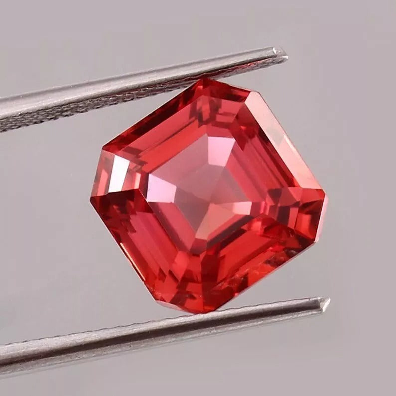 Padparadscha Sapphire gemstone held between tweezers on a gray background