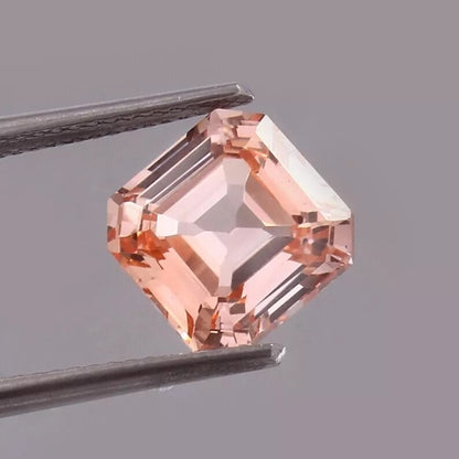 asscher Padparadscha Sapphire – Peach gemstone held between tweezers on a gray background