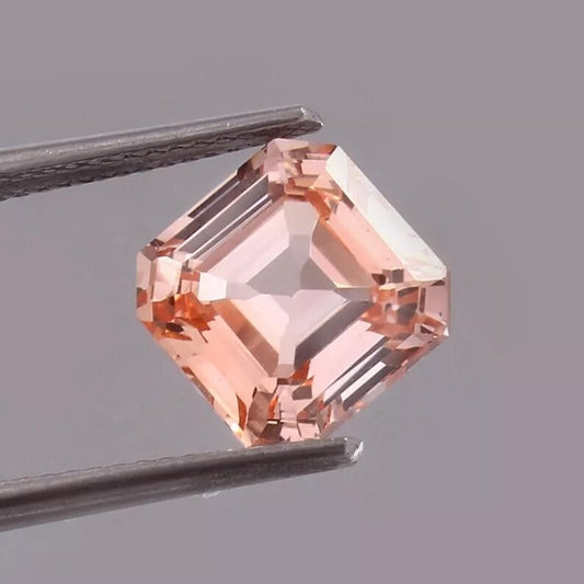asscher Padparadscha Sapphire – Peach gemstone held between tweezers on a gray background