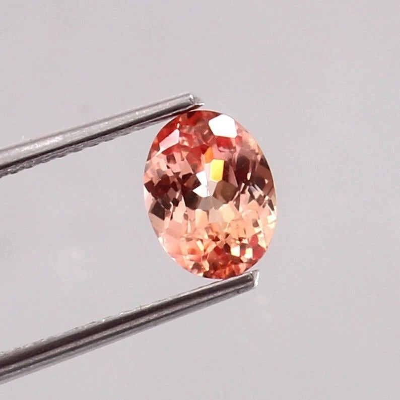 Padparadscha Sapphire gemstone held by tweezers on a light gray background