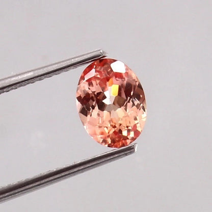 Padparadscha Sapphire gemstone held by tweezers on a light gray background