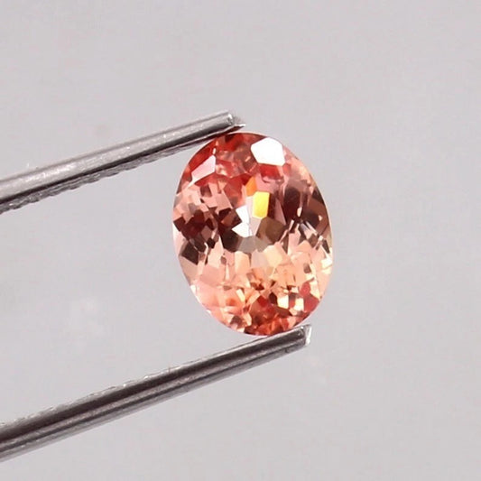 Padparadscha Sapphire gemstone held by tweezers on a light gray background