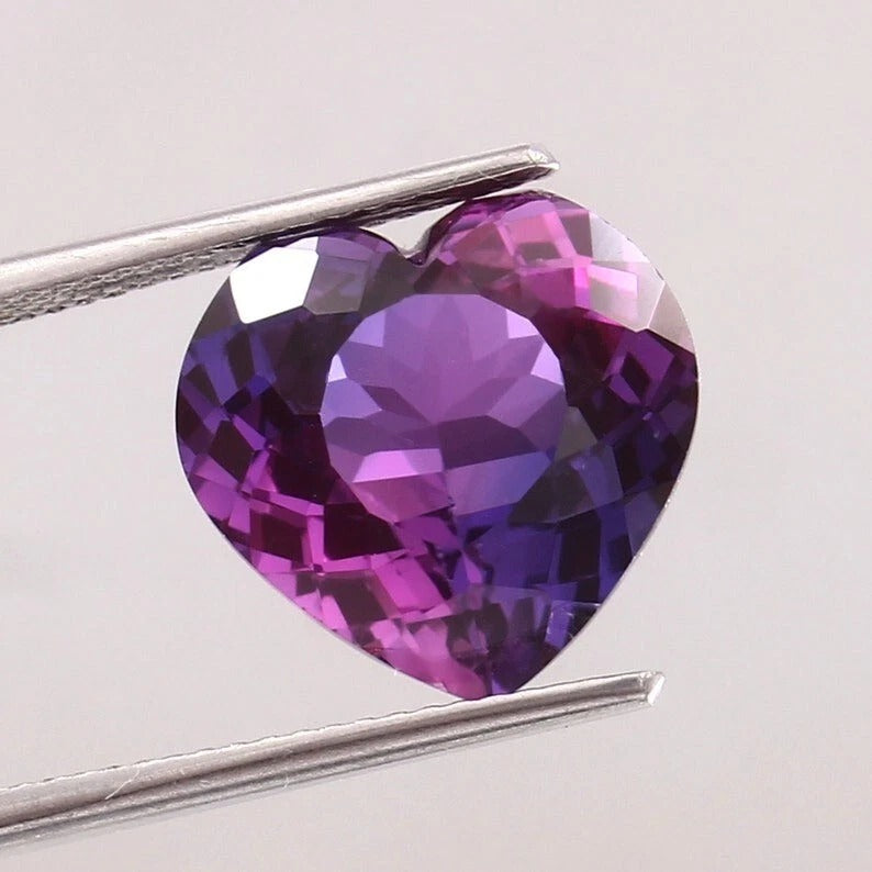 Heart-shaped Purple-Pink Sapphire gemstone held by tweezers on a light gray background