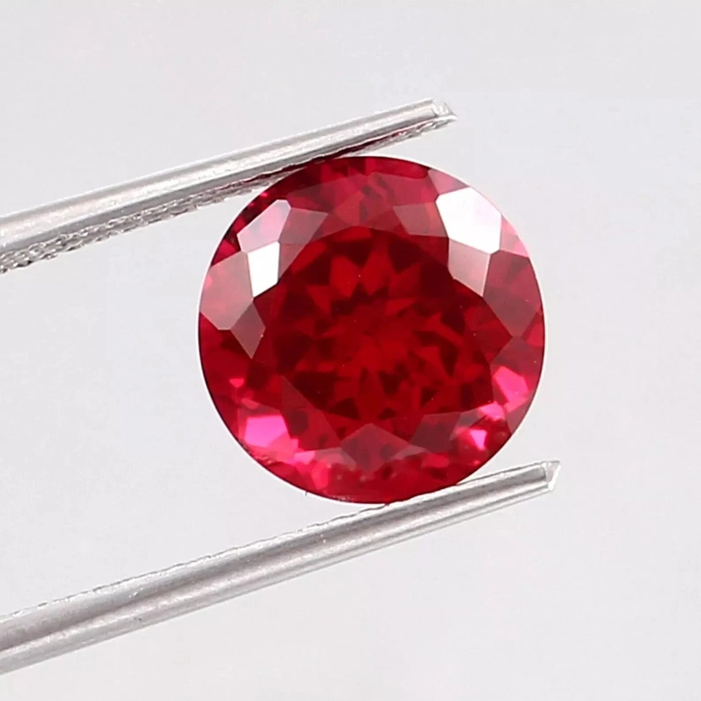 Round red Mozambique Ruby gemstone held between tweezers on a light gray background