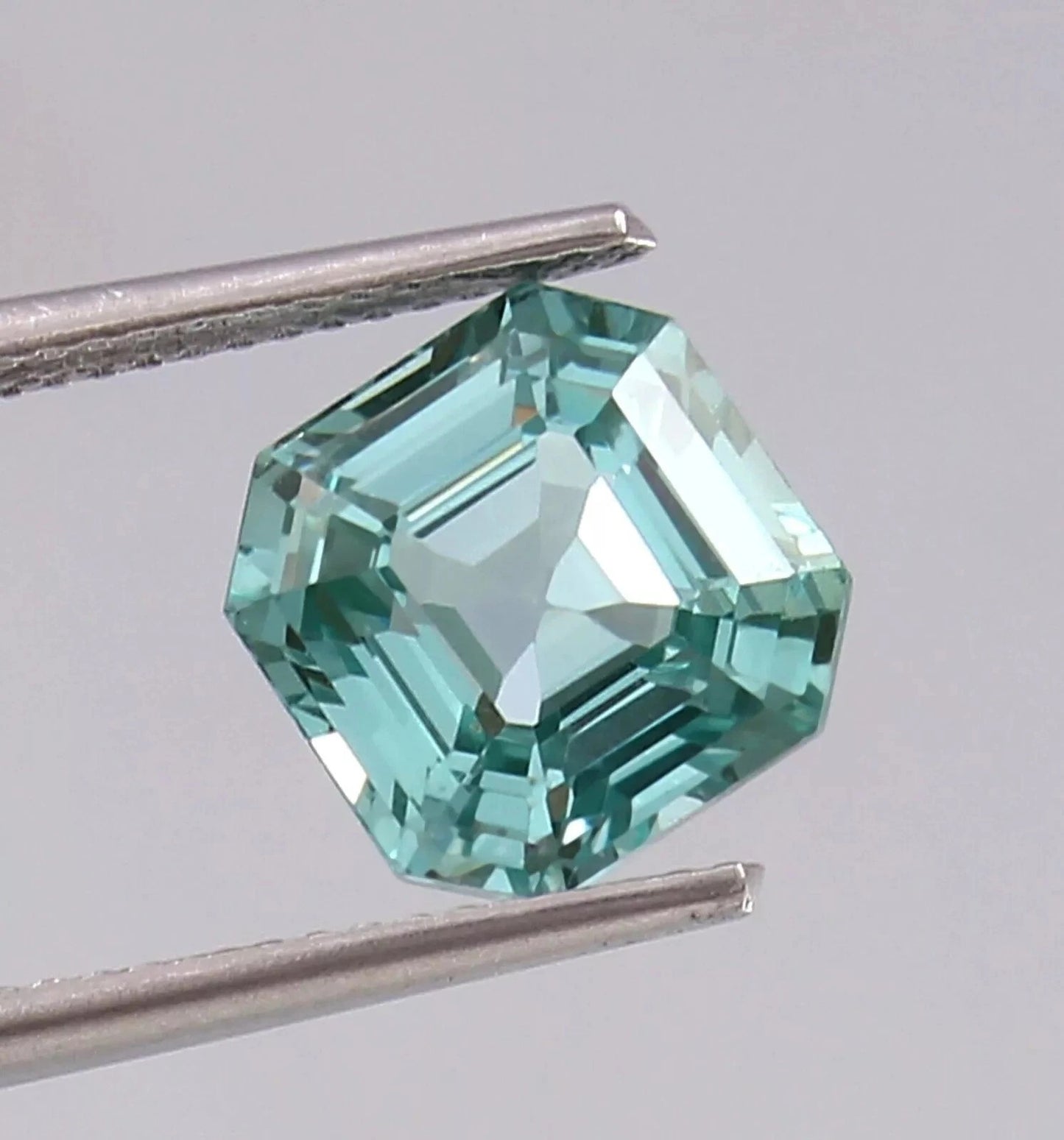 Green Paraiba Tourmaline gemstone held by tweezers against a gray background