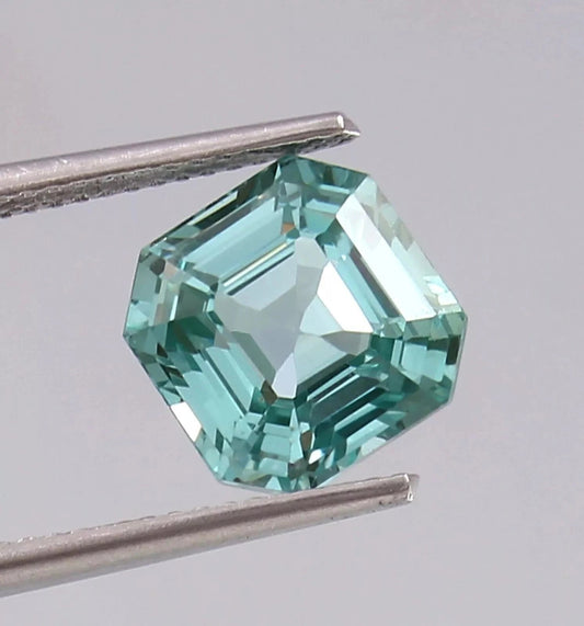Green Paraiba Tourmaline gemstone held by tweezers against a gray background