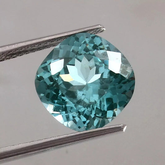 Teal Spinel  gemstone held by tweezers on a gray background